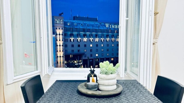 A hotel room with a view of a building with 'WIMBLED' signage, a table with a placemat, two chairs, and a small potted plant.