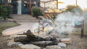 A fire pit with rocks and logs, surrounded by a wooden bench and a chair, with a building in the background.