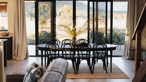 A dining area with a round table and chairs, a vase with yellow flowers, and a view of a natural landscape through large windows.