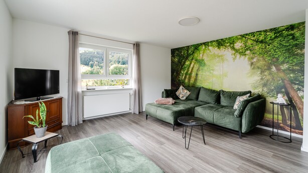 A living room with a green sofa, a television, a bench, and a large forest mural on the wall. - Waldrose Apartment with Mountain View, Private Terrace, and Wi-Fi (Oberharmersbach)