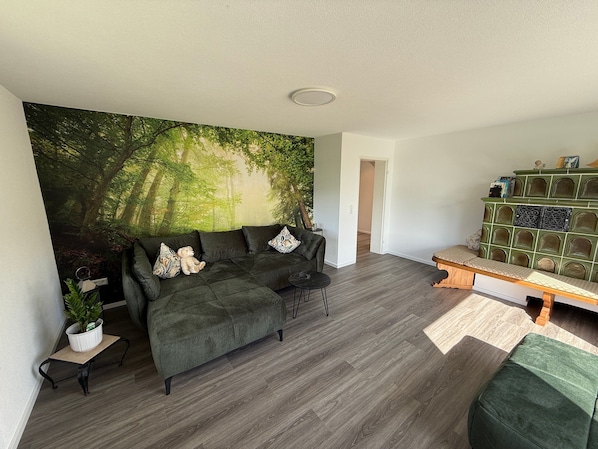 Un salon avec un canapé vert, une petite table sur laquelle est posée une plante, une bibliothèque et une grande fresque représentant une forêt sur le mur.