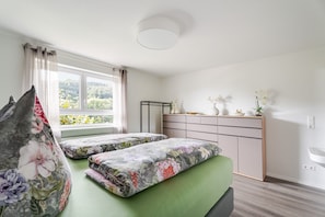 Ein Schlafzimmer mit zwei Betten, einem Fenster mit Vorhängen, einem Nachttisch mit Dekoration und einer Kommode mit Schubladen.