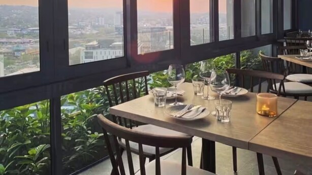 Un restaurante con una mesa para dos, amplios ventanales que ofrecen vista a la ciudad y plantas de interior.
