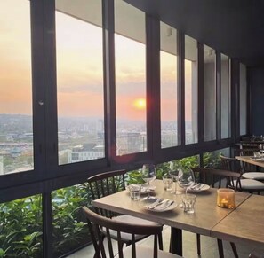 Un restaurant avec une table dressée pour deux personnes, de grandes fenêtres donnant sur la ville et des plantes d’intérieur.