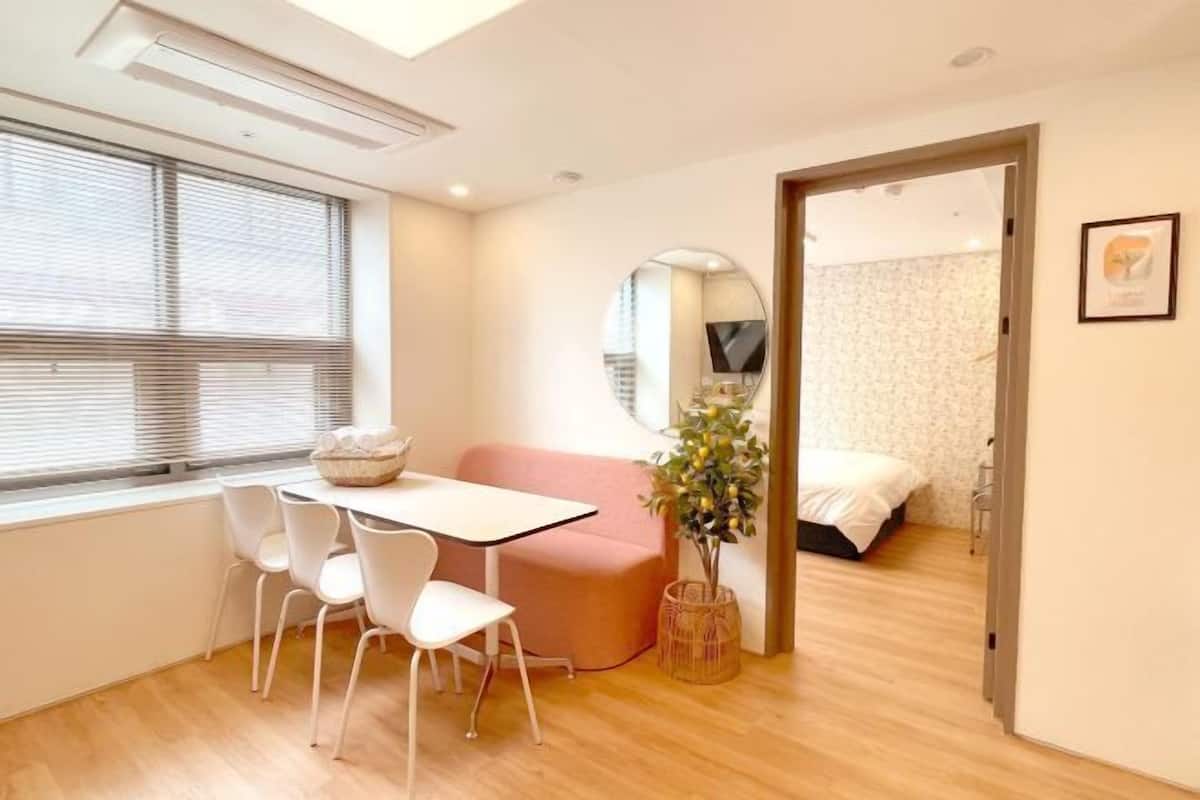 Een moderne kamer met een eettafel, stoelen, een roze bank, een kleine plant, een bed en een spiegel.
