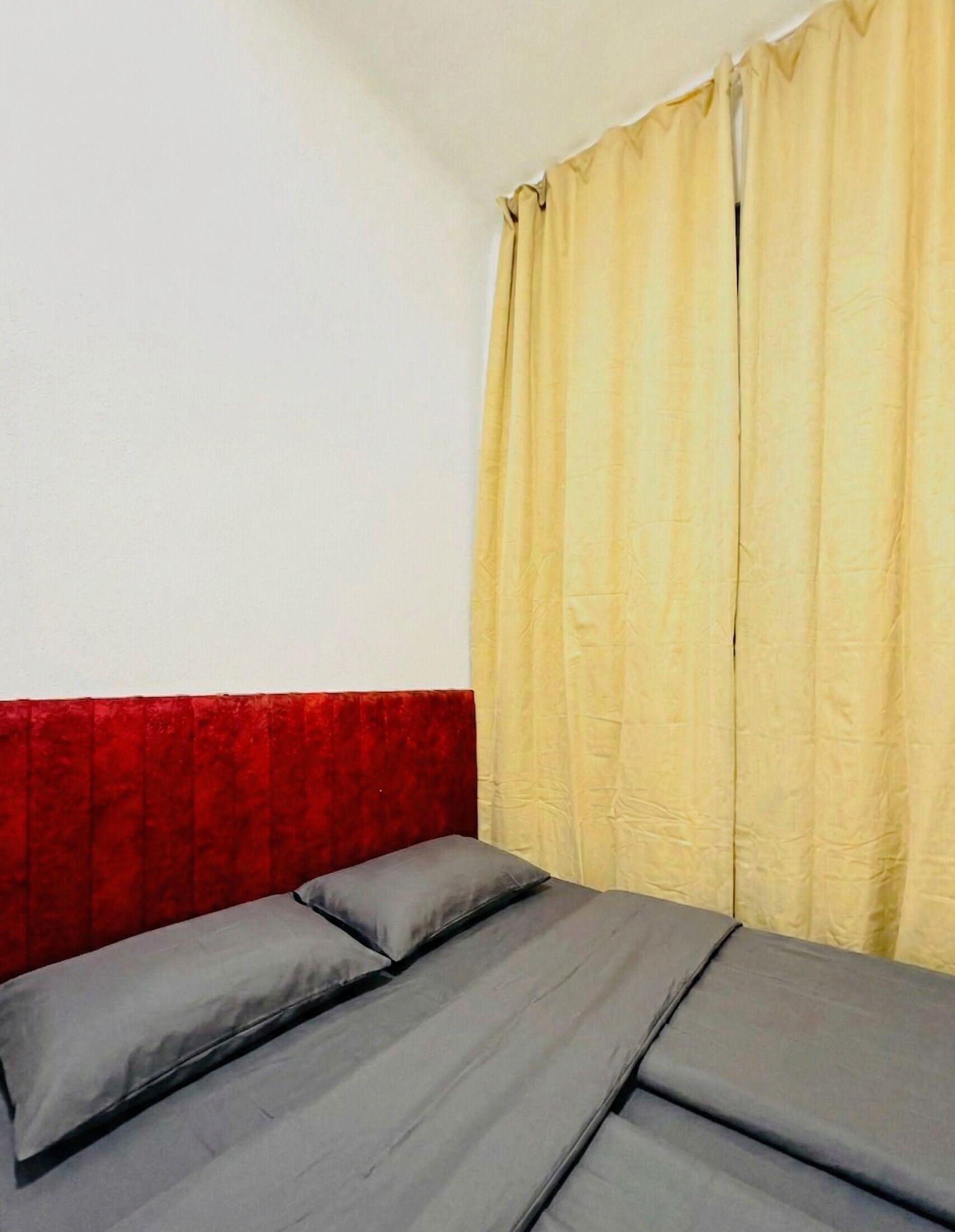 Una cama con ropa de cama gris y cabecera roja en una habitación con pared blanca lisa y cortinas amarillas.