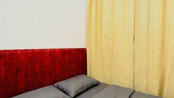 Una cama con ropa de cama gris y cabecera roja en una habitación con pared blanca lisa y cortinas amarillas.