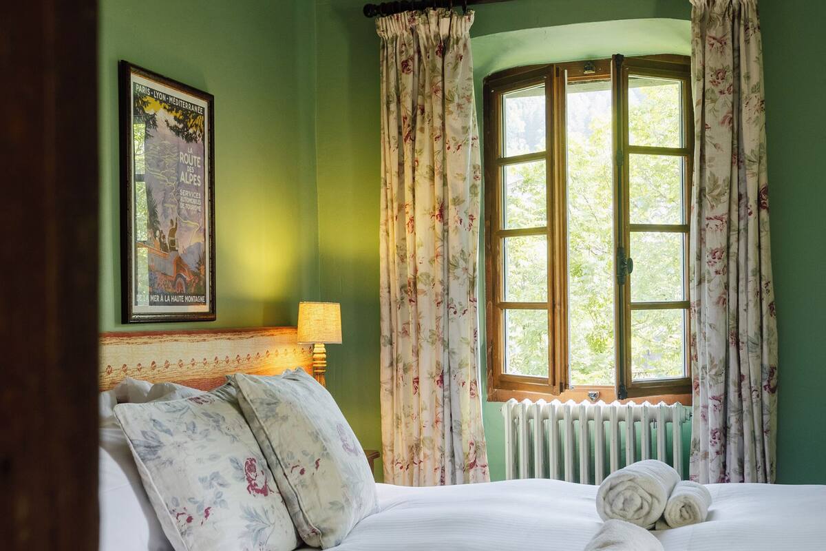 Una camera da letto con un letto, tende floreali, una finestra che si affaccia sul verde, un termosifone e un poster incorniciato al muro.
