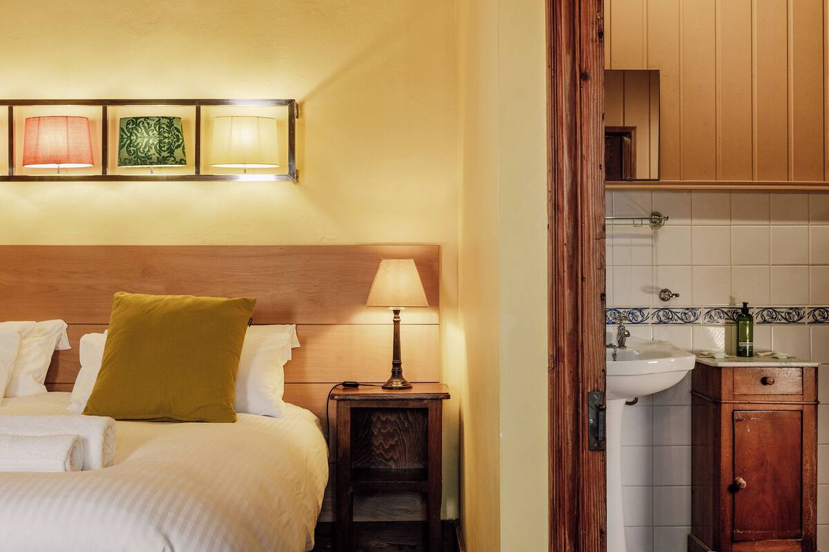 Una camera d'albergo con un letto, un comodino con una lampada, una testiera in legno e vista sul bagno attraverso una porta aperta.
