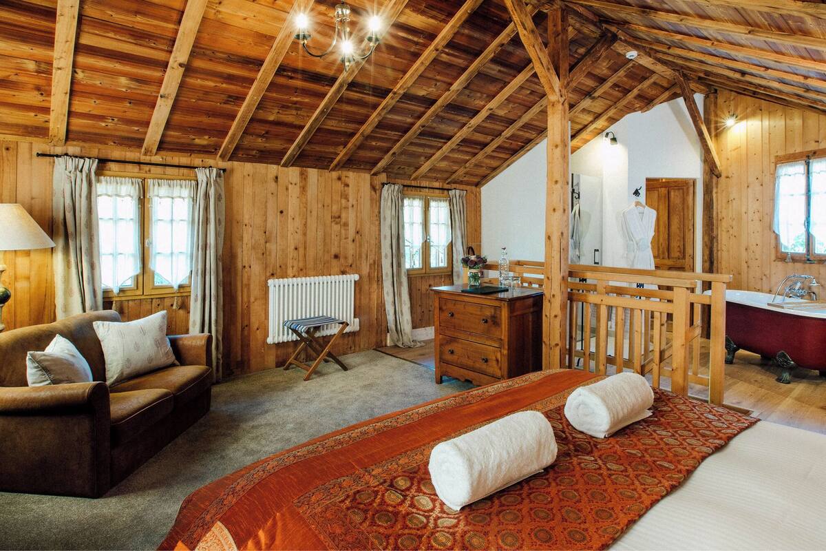 Interno di una baita in legno con un letto, un divano, una cassettiera, un termosifone, una vasca da bagno e una finestra.
