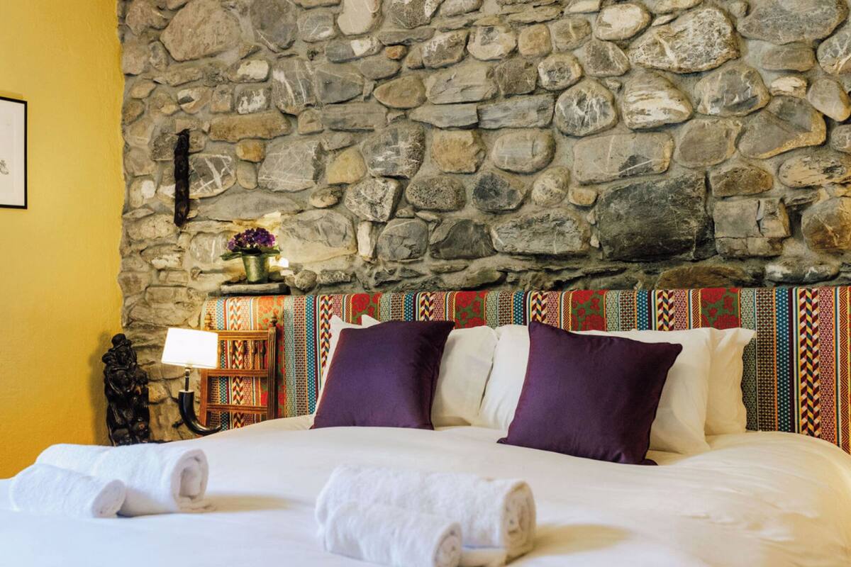 Un letto con lenzuola bianche e cuscini viola, una parete in pietra e una testiera colorata con motivi decorativi.