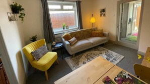 Ein Wohnzimmer mit einer Couch, einem gelben Sessel, einem Holztisch, einem gemusterten Teppich und einem Fenster mit Vorhängen.