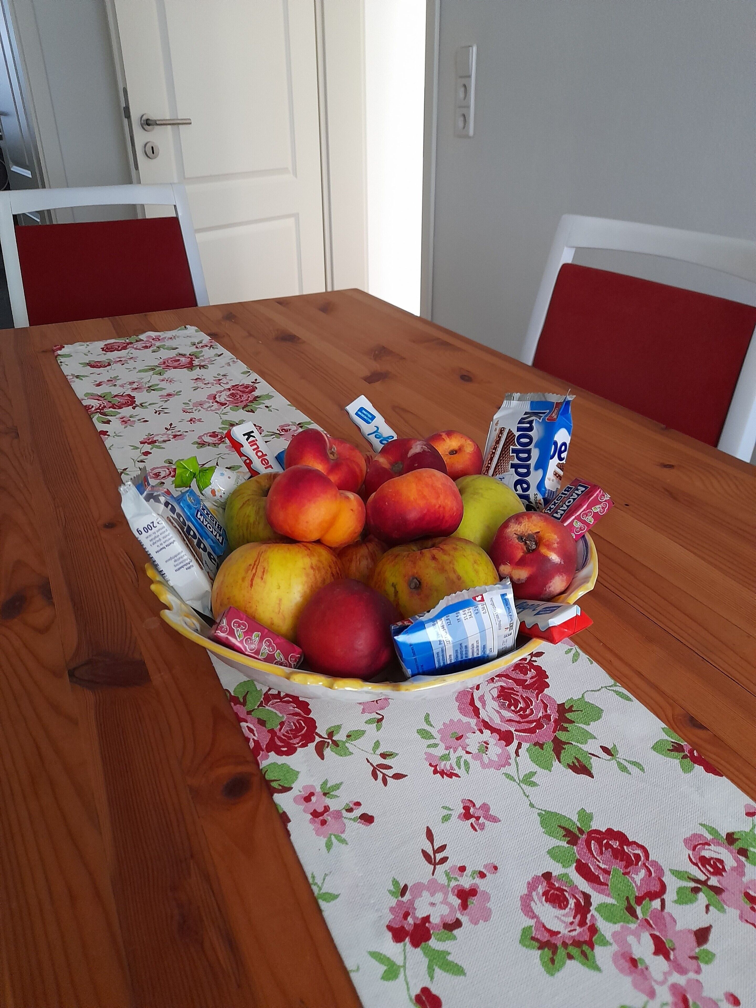 A wooden dining table with a floral tablecloth, a fruit basket, and packaged snacks.