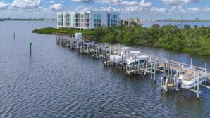 A waterfront property with a dock extending into the water, a building with multiple balconies, and a clear sky with clouds. - Sunset Views, Sand Between Your Toes-Your Winter Escape Awaits! (Englewood)