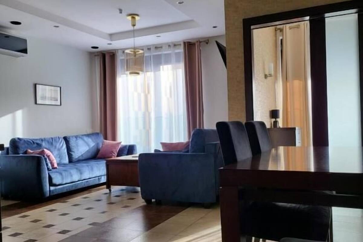 A modern living room with a sofa, a dining table with chairs, and a large window with curtains.