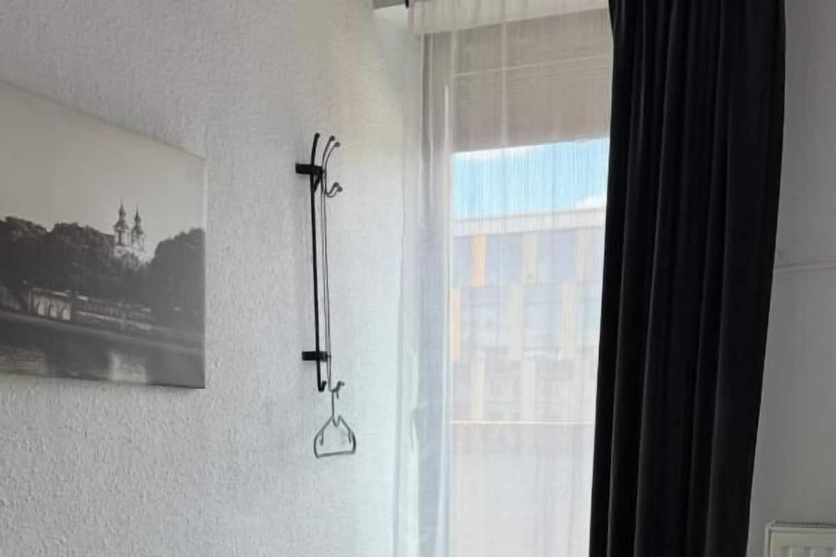 A neatly made bed with a white pillow and a folded blanket, a black curtain partially drawn, and a framed picture on the wall.