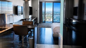A modern hotel room with a kitchen area, a dining table, a sofa, and a large window offering a city view.