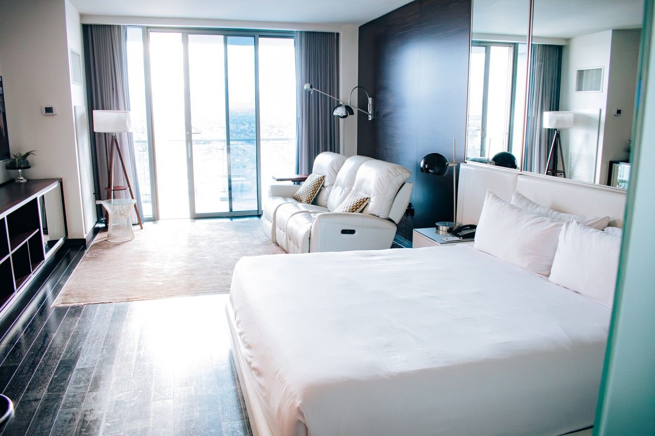 A modern hotel room with a large bed, a seating area with a sofa, a desk with a lamp, and a large window with a view.
