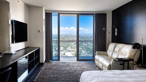 A modern hotel room with a large bed, a flat-screen TV, a sofa, and a view of the cityscape through floor-to-ceiling windows.