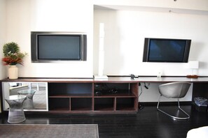 A modern hotel room with a dark wood desk, two flat-screen TVs mounted on the wall, a small potted plant, and a chair.