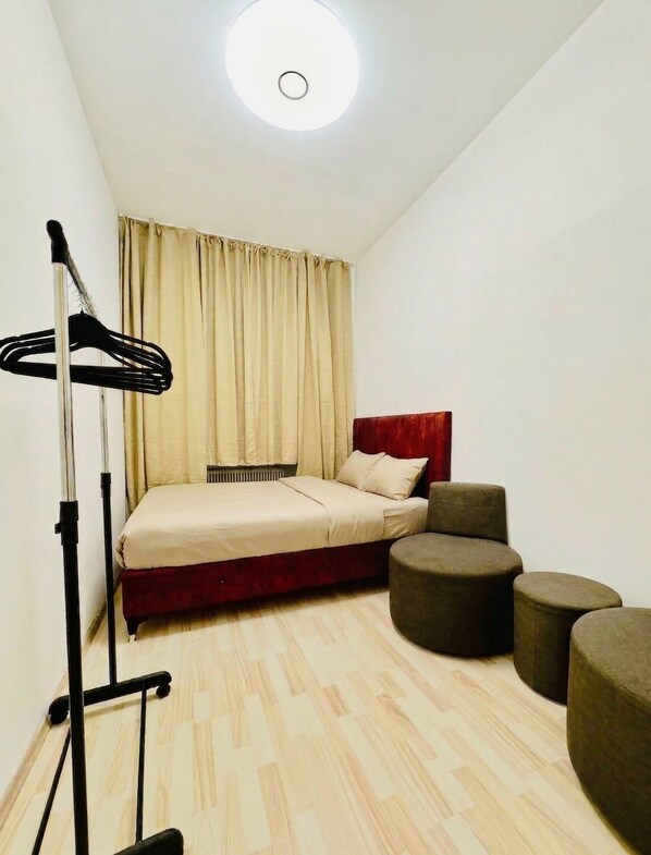 A compact bedroom with a bed, a standing lamp, a radiator, and a small seating area with ottomans.