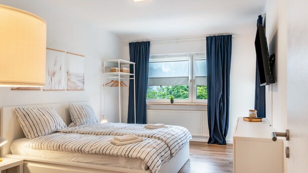 Une chambre à coucher avec un lit, une télévision, une fenêtre avec des rideaux et une étagère avec divers objets.