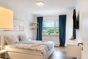 Un dormitorio con una cama, un televisor, una ventana con cortinas y una estantería con objetos.