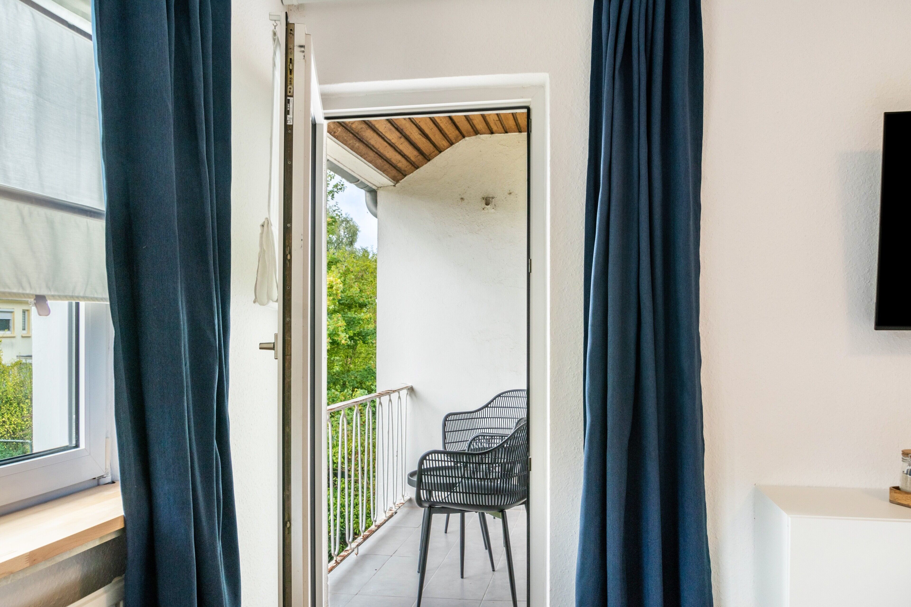 Eine offene Tür führt zu einem Balkon mit zwei Stühlen und einer Holzbalkendecke.
