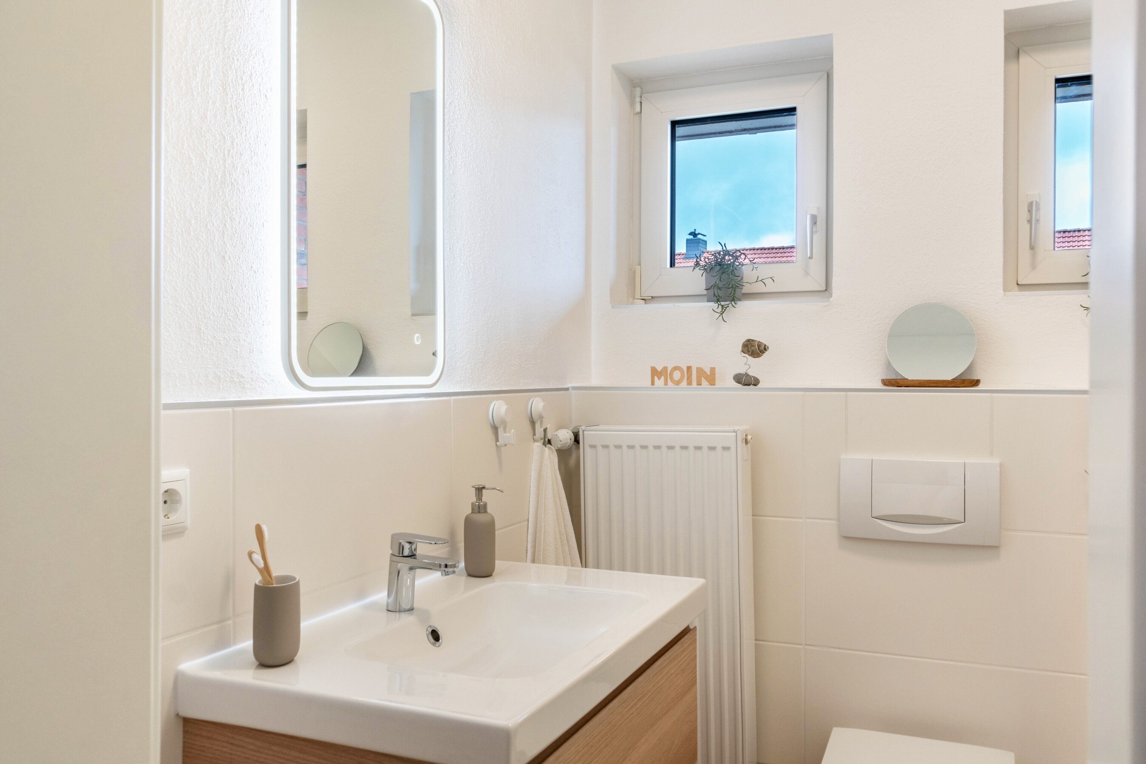 Une salle de bain avec un lavabo blanc, un miroir et une fenêtre donnant sur des immeubles.