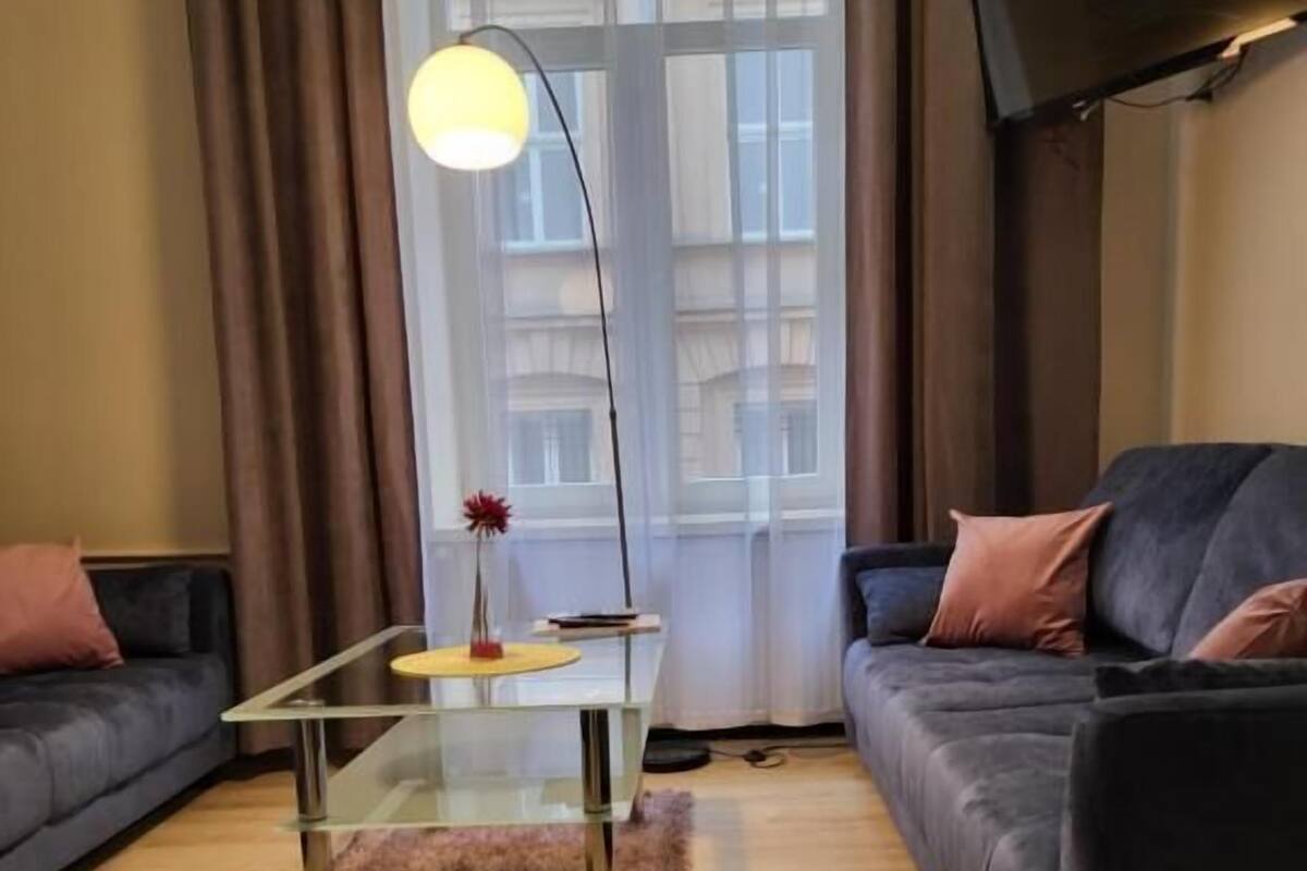 A modern living room with a glass coffee table, a sofa, a rug, and a large window with curtains.