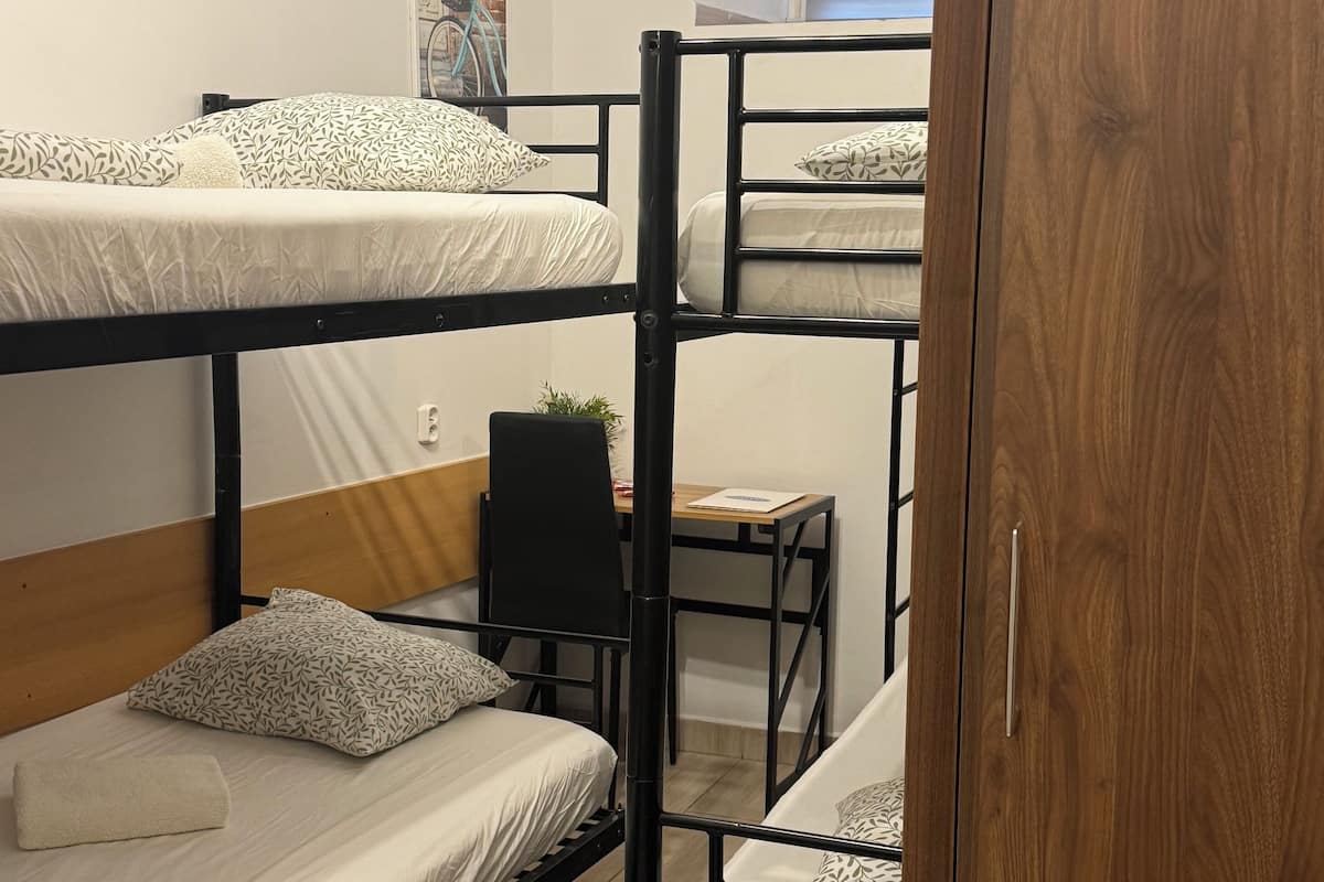 A bunk bed room with a desk and chair, a wooden wardrobe, and a window with blinds.