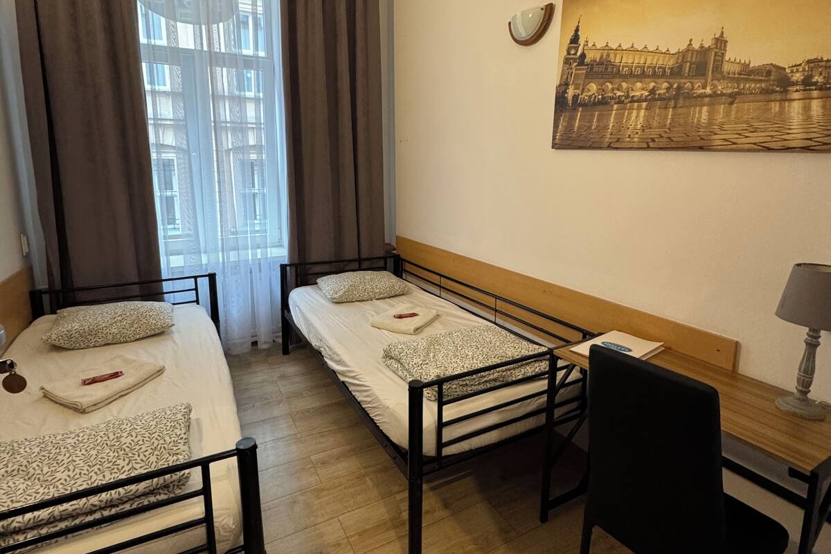 A room with two single beds, a desk, a chair, a lamp, and a large window with curtains.