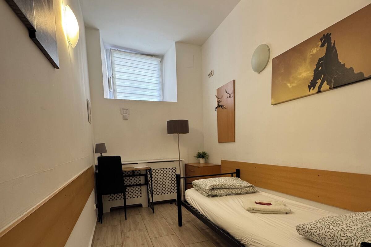 A small, well-lit room with a bed, a desk, and a window.