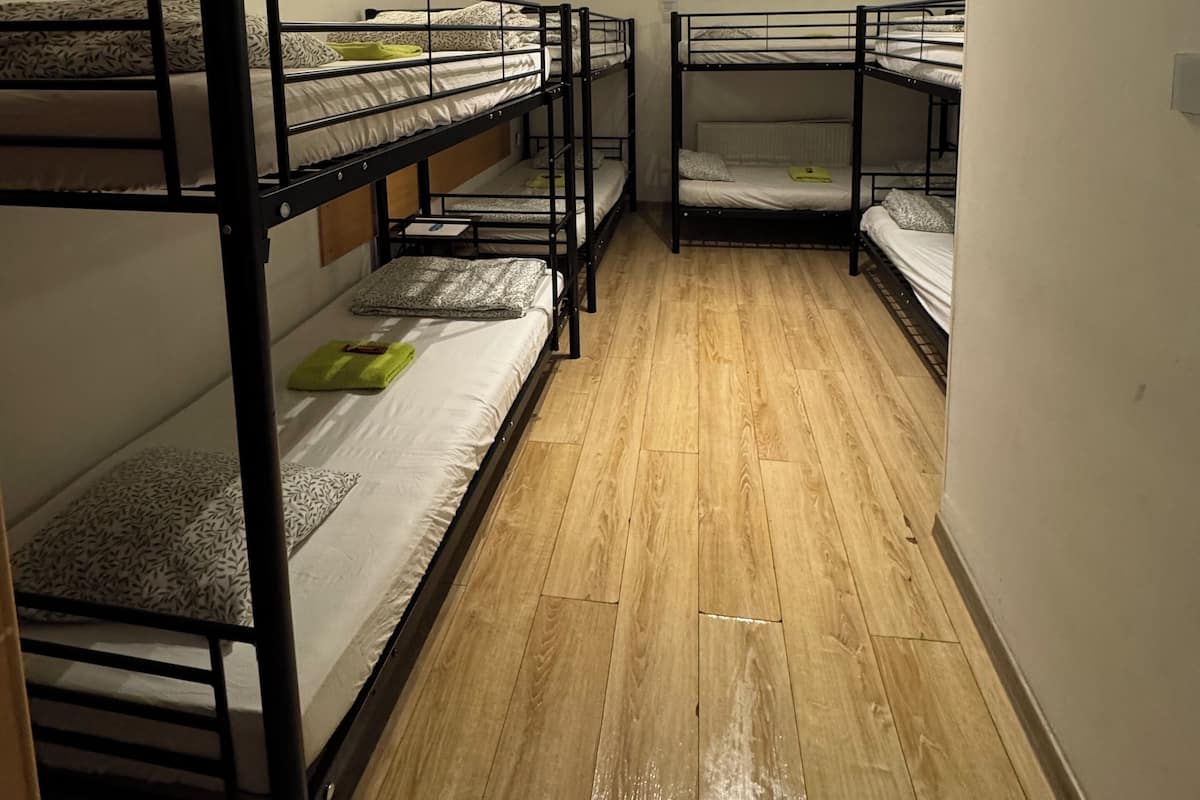 A dormitory room with bunk beds, wooden flooring, a ceiling fan, and a window with blinds.
