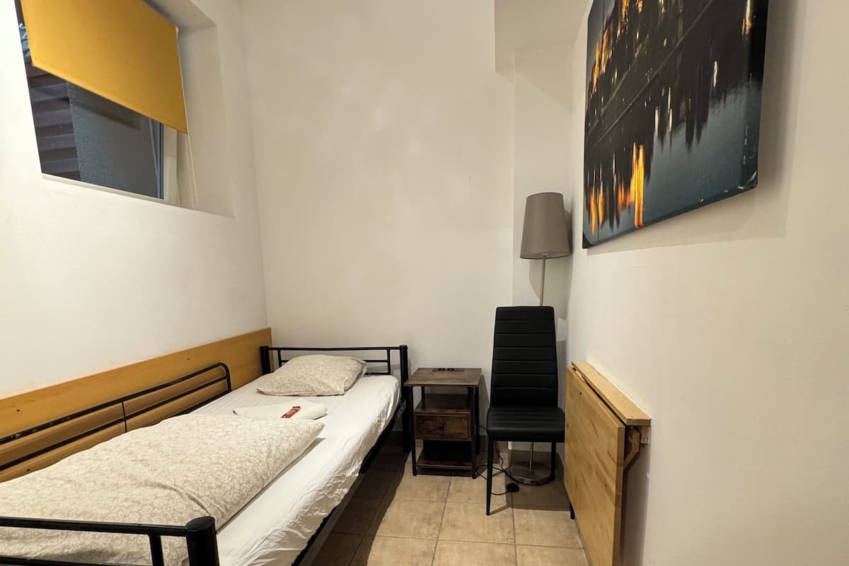 A small, narrow room with a single bed, a nightstand, a chair, and a radiator.