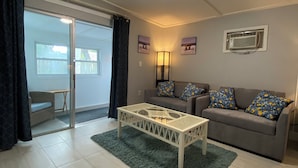 Ein modernes Wohnzimmer mit einem grauen Sofa, gemusterten Kissen, einem Glastisch und einer Glastür, die zu einem Außenbereich führt.
