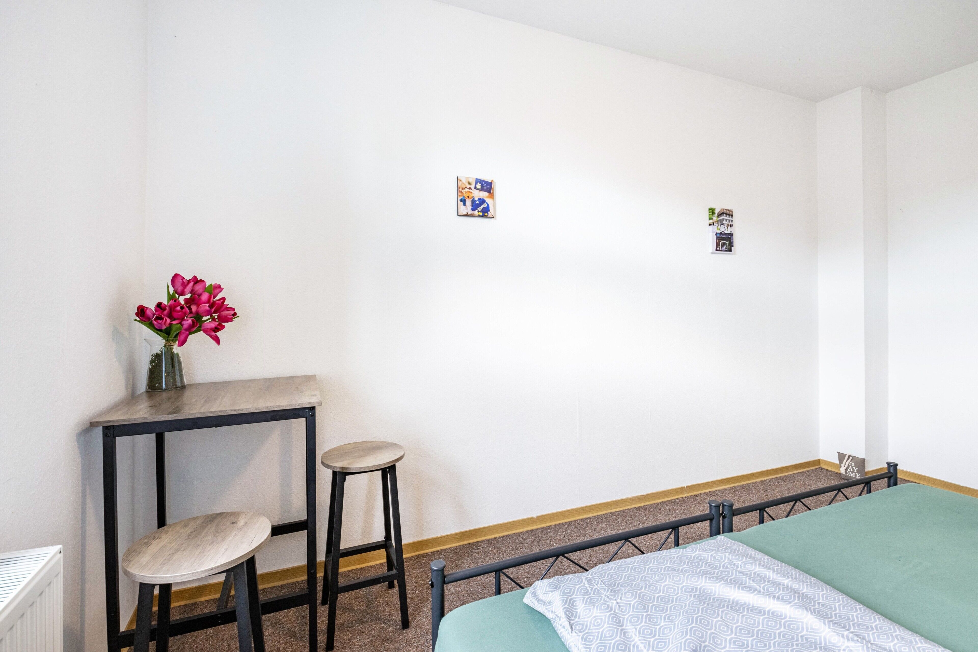 Una habitació amb un llit, una taula petita amb un ram de flors, dos tamborets i un radiador.