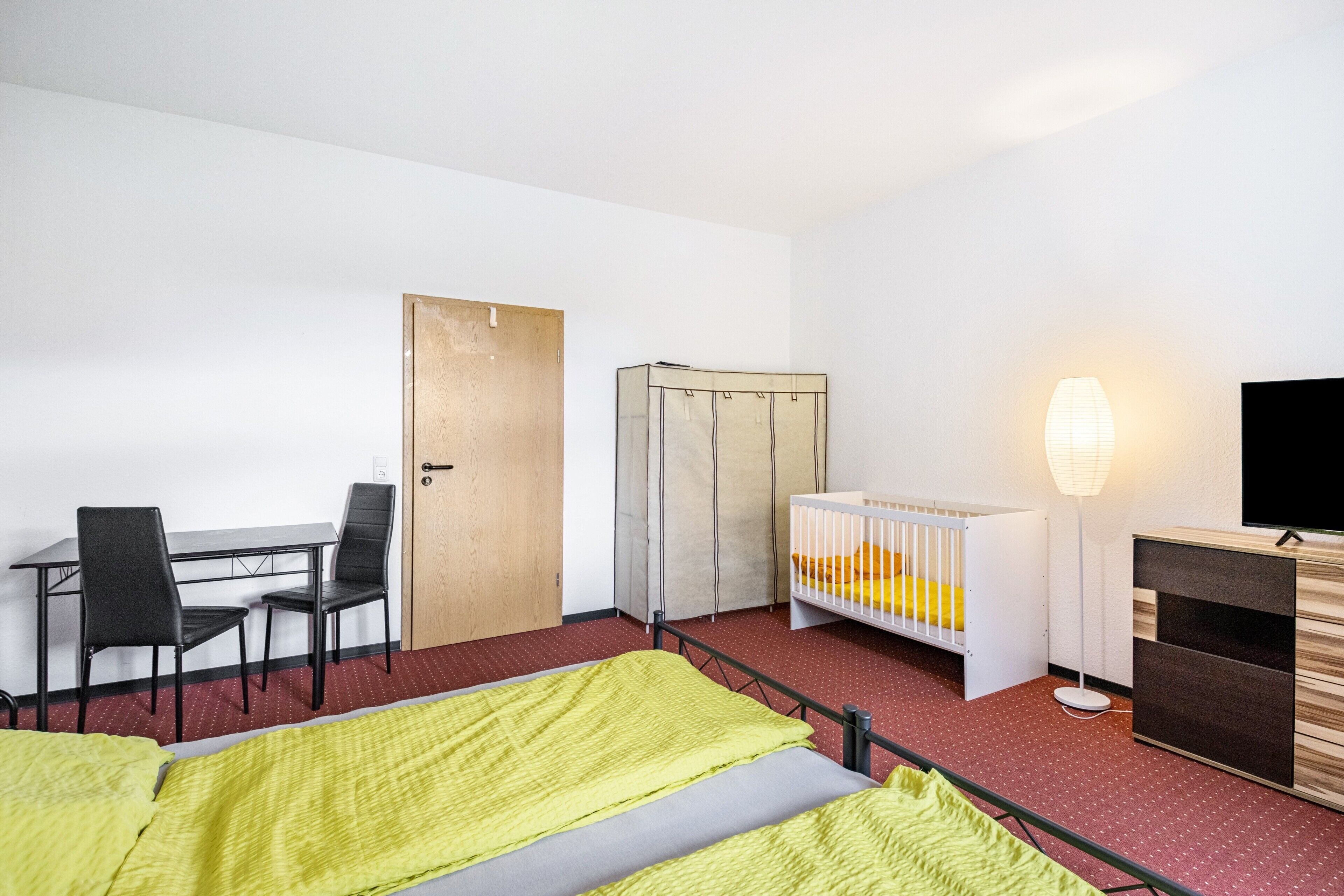 A hotel room with a bed, a desk with chairs, a crib, a TV, and a wardrobe.