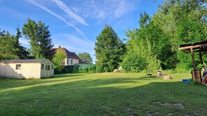 Un jardin avec une pelouse, un petit abri de jardin, une balançoire en bois et une maison avec un toit rouge.