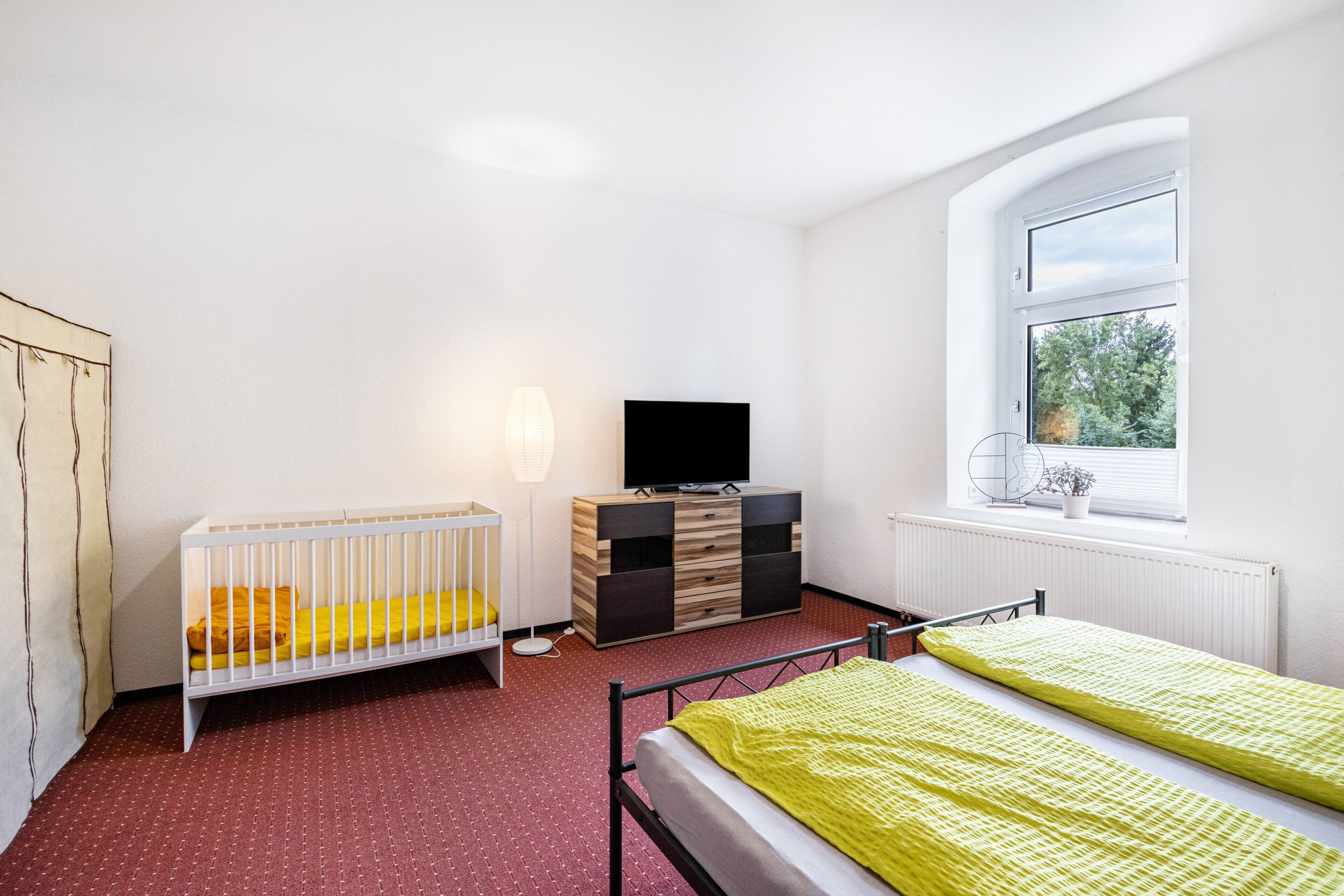 Ein Zimmer mit einem Kinderbett, einem Bett, einem Fernseher, einer Kommode und einem Fenster mit Aussicht.