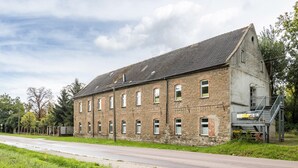 Un grand bâtiment historique en pierre, doté de nombreuses fenêtres et d’un toit en pente, situé au bord d’une route asphaltée.
