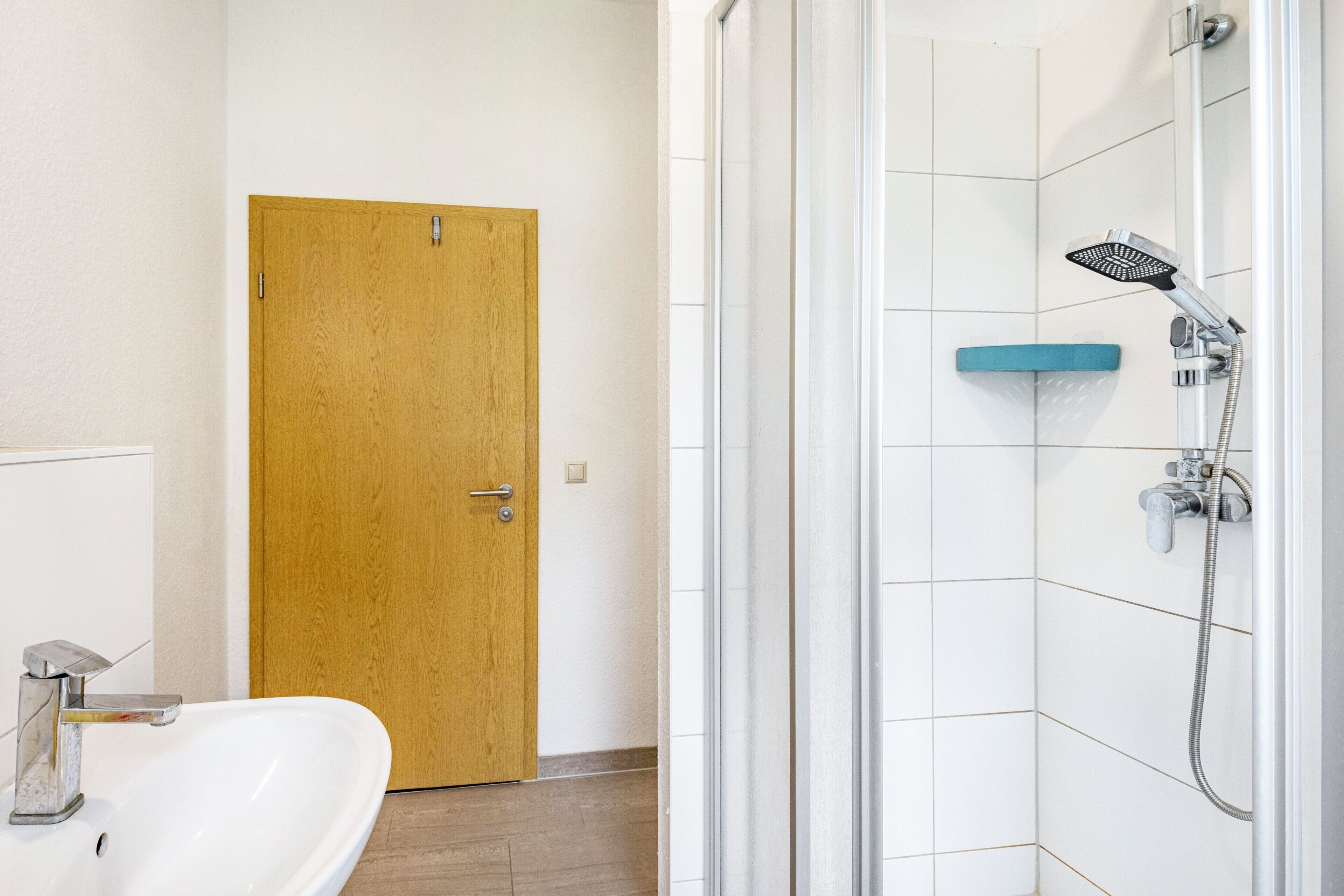 Une salle de bain avec une porte en bois, un lavabo blanc et une douche équipée d’un pommeau de douche à main.