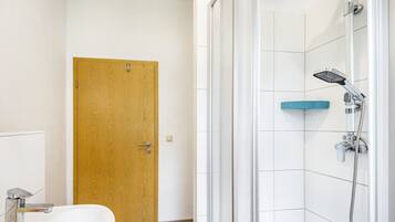 Une salle de bain avec une porte en bois, un lavabo blanc et une douche équipée d’un pommeau de douche à main.