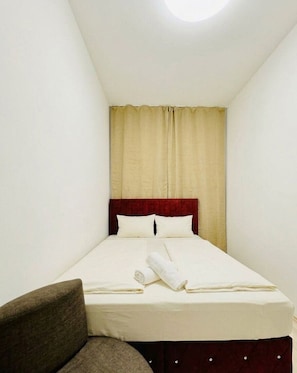 A small, single-bed room with a red headboard, a brown armchair, and a window with beige curtains.