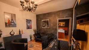 Een moderne woonkamer met een leren bank, een glazen salontafel en een kroonluchter. Er hangen ingelijste foto's aan de muren, een tafeltje met flessen, en een bed op de achtergrond.