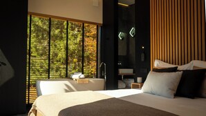 Een moderne hotelkamer met een groot bed, een zithoek met een stoel en tafel, een groot raam met uitzicht op bomen, en een wand met houten accenten.