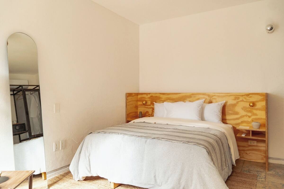 Quarto com cabeceira de madeira, cama com roupa de cama branca, mesa de cabeceira com abajur e espelho na parede.