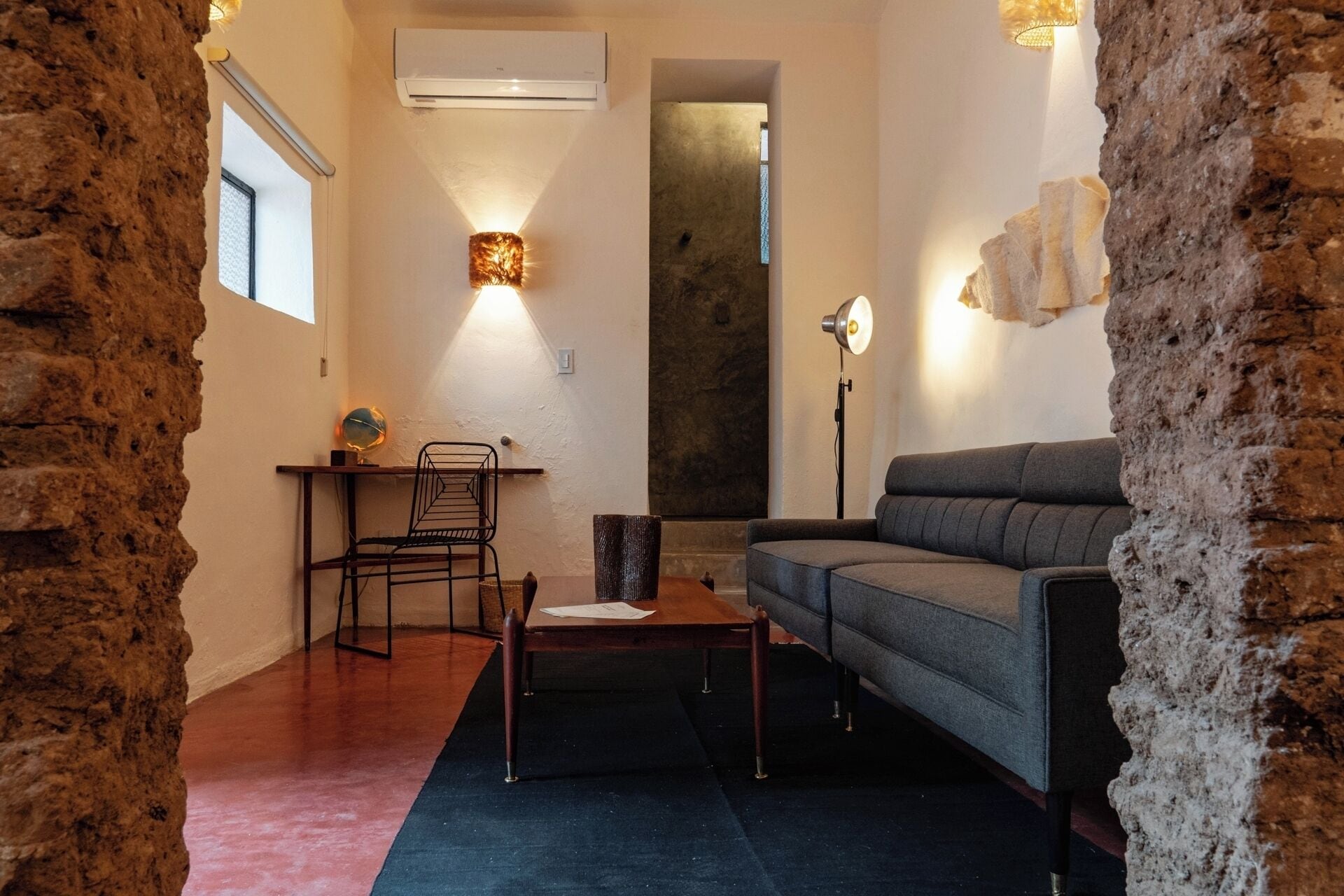 Un salon confortable avec un canapé, un fauteuil, une petite table et un bureau sur lequel se trouve un globe terrestre. Une grande fenêtre et un appareil de climatisation sont fixés au mur.
