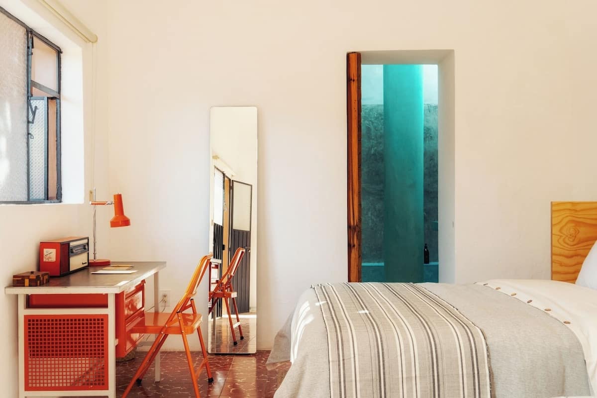 Quarto com cama, escrivaninha, cadeira, espelho e janela.