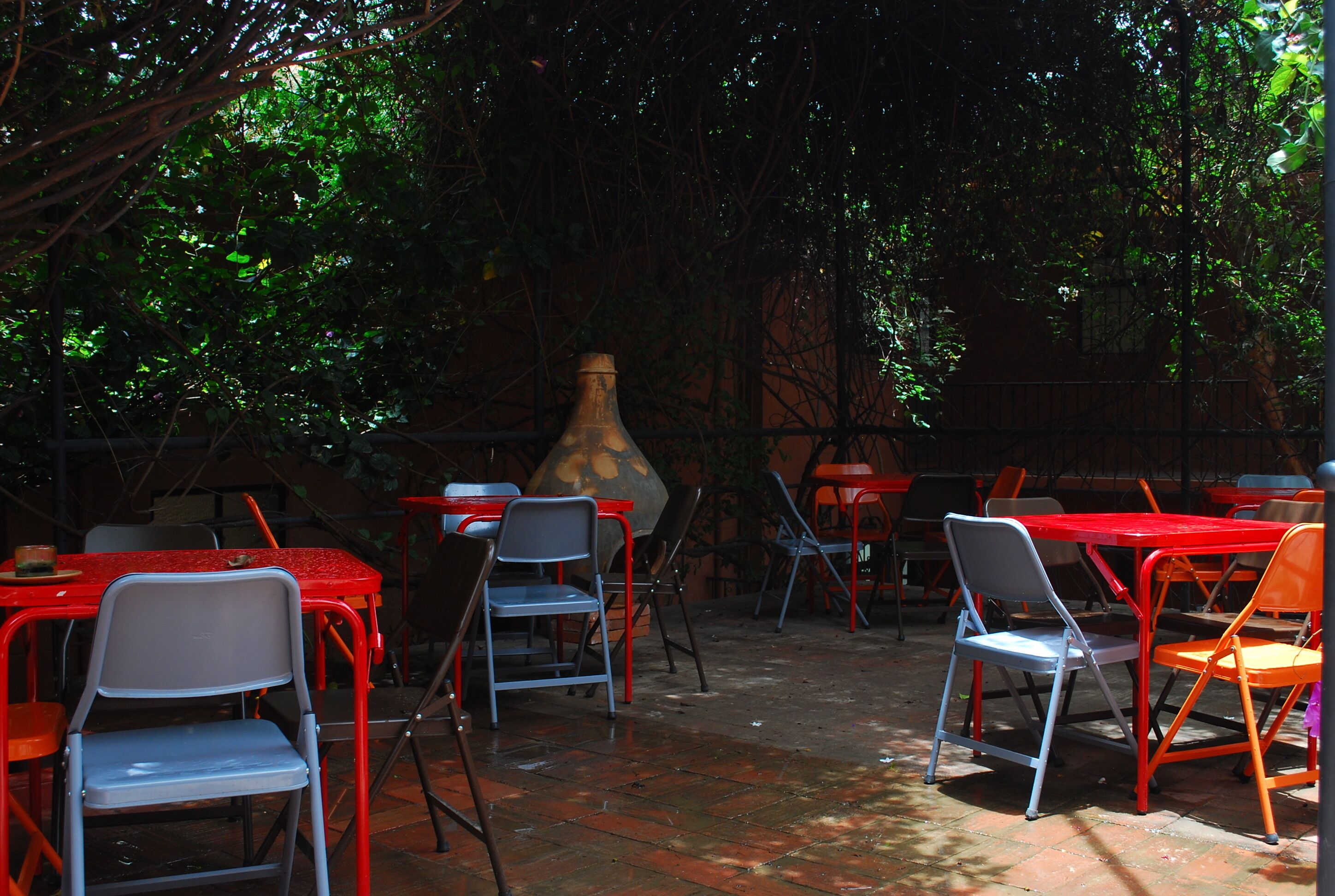 Un patio al aire libre con mesas y sillas de metal rojo y gris, rodeado de follaje.